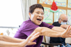 紫色のシャツを着た男性が両腕を広げて楽しく体操・運動をしている風景。背景に他の入居者も見える。